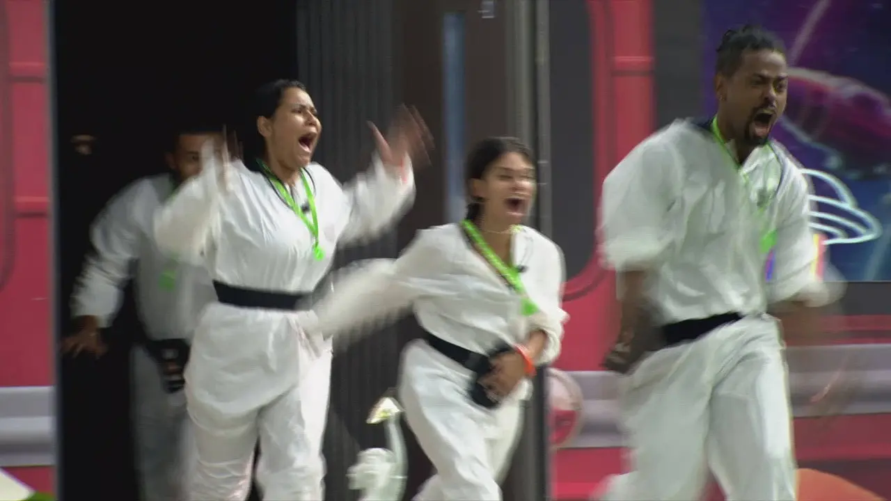 Participantes do BBB 26 correm e gritam entusiasmados ao entrar na casa, vestindo macacões brancos com faixas pretas e cordões verdes. A cena captura um momento de intensa euforia e movimento, com os integrantes saindo do quarto branco para o cenário da casa do reality.