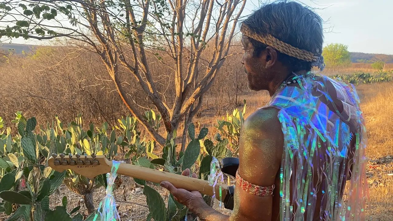 Um homem de perfil segura uma guitarra em meio a uma paisagem árida repleta de cactos e árvores secas sob a luz do entardecer. Ele utiliza adornos tradicionais, como uma faixa na cabeça e no braço, combinados com uma capa de fitas iridescentes que refletem cores vibrantes, unindo elementos ancestrais e contemporâneos.