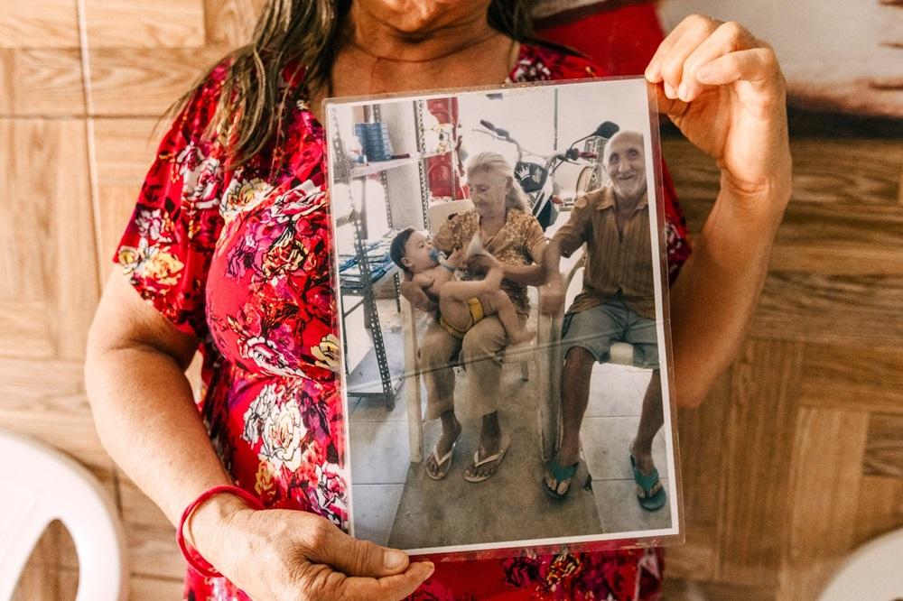 Na imagem, uma mulher veste um vestido floral vermelho e segura, na altura do peito, uma fotografia vertical protegida por um plástico transparente. Na foto impressa, o casal de idosos, Seu Francisco e Dona Luzia, aparece em um momento descontraído e sorridente; ela está sentada com uma criança pequena no colo, enquanto ele está sentado ao lado em uma cadeira de plástico. Ao fundo da fotografia, nota-se uma motocicleta e prateleiras. A mulher que segura a foto está em um ambiente com paredes que imitam madeira clara, criando um contraste vibrante entre o vermelho de sua roupa e a imagem antiga que ela preserva.