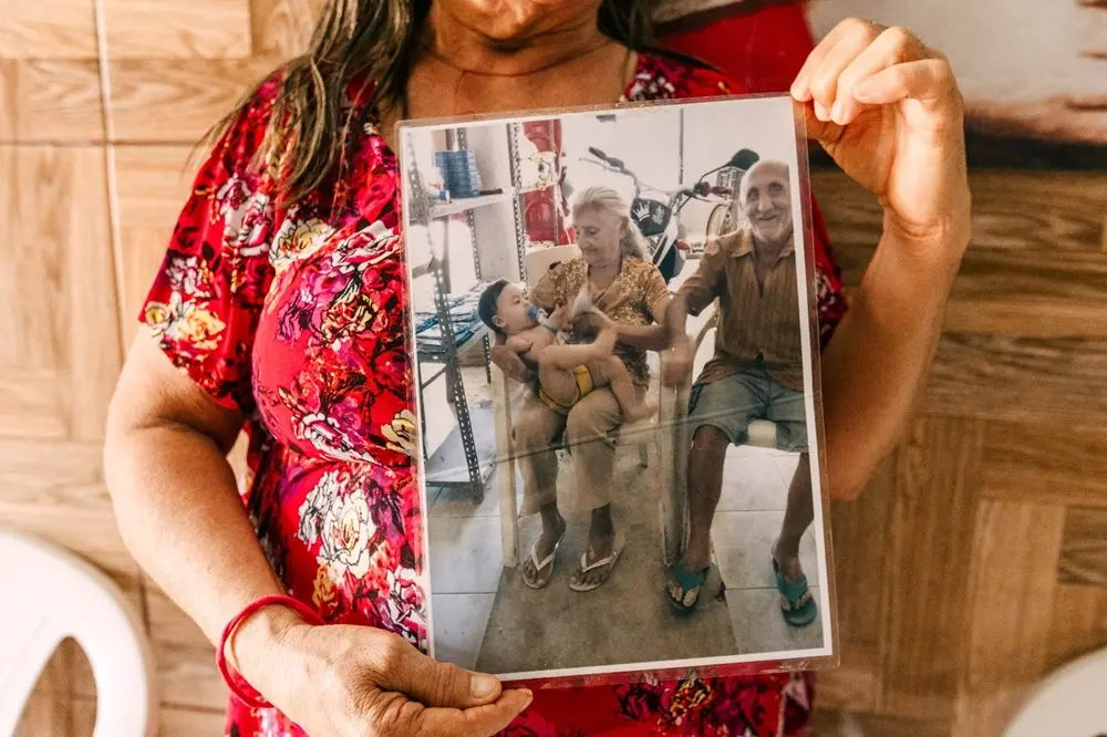 Na imagem, uma mulher veste um vestido floral vermelho e segura, na altura do peito, uma fotografia vertical protegida por um plástico transparente. Na foto impressa, o casal de idosos, Seu Francisco e Dona Luzia, aparece em um momento descontraído e sorridente; ela está sentada com uma criança pequena no colo, enquanto ele está sentado ao lado em uma cadeira de plástico. Ao fundo da fotografia, nota-se uma motocicleta e prateleiras. A mulher que segura a foto está em um ambiente com paredes que imitam madeira clara, criando um contraste vibrante entre o vermelho de sua roupa e a imagem antiga que ela preserva.