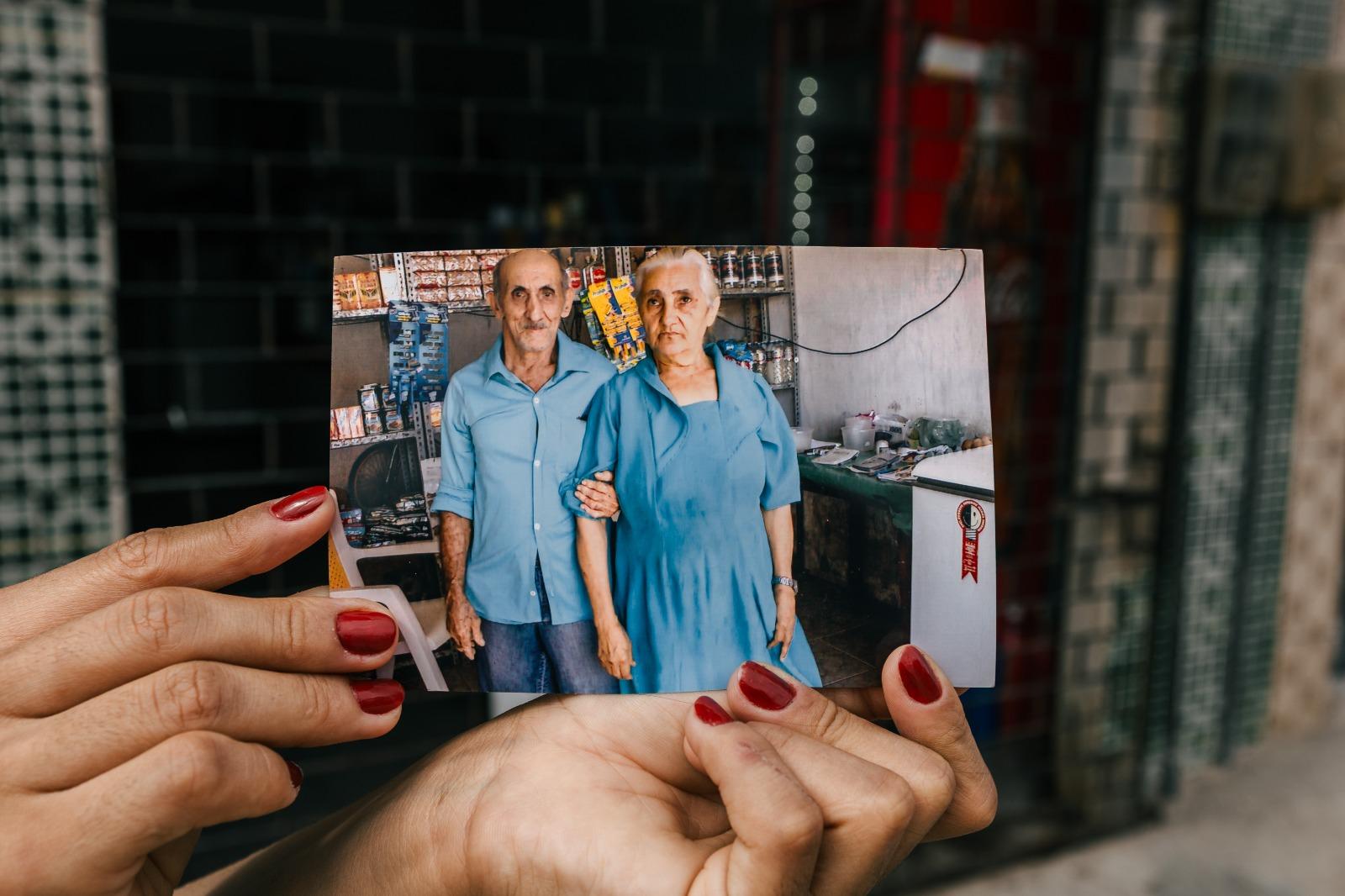 Na imagem, uma pessoa com unhas pintadas de vermelho segura uma fotografia horizontal em frente a uma parede de azulejos escuros e uma fachada comercial. A fotografia mostra o casal de idosos, Seu Francisco e Dona Luzia, em pé dentro de sua mercearia. Ambos vestem roupas azuis; Dona Luzia está com a mão no braço de Seu Francisco. O fundo da fotografia revela prateleiras com mercadorias e um freezer branco. A mão que segura a foto está em destaque no primeiro plano, criando um contraste entre a textura da pele e a imagem estática do passado.