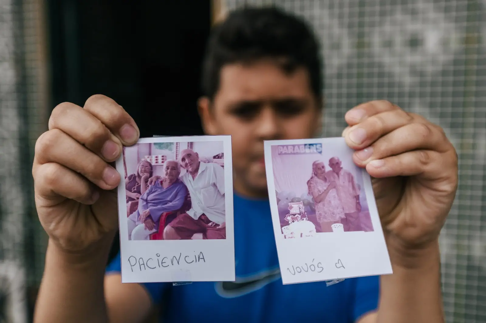 Na imagem, um jovem, com o rosto levemente fora de foco ao fundo, segura duas fotos em estilo Polaroid em direção à câmera. Na mão direita, a foto mostra o casal de idosos, Seu Francisco e Dona Luzia, sentados e sorrindo; abaixo da imagem está escrita à mão a palavra 
