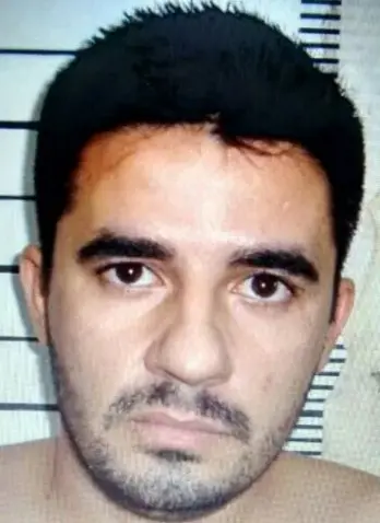 Retrato de um homem de pele clara, cabelo preto curto, barba por fazer e expressão séria, com fundo branco com linhas horizontais.