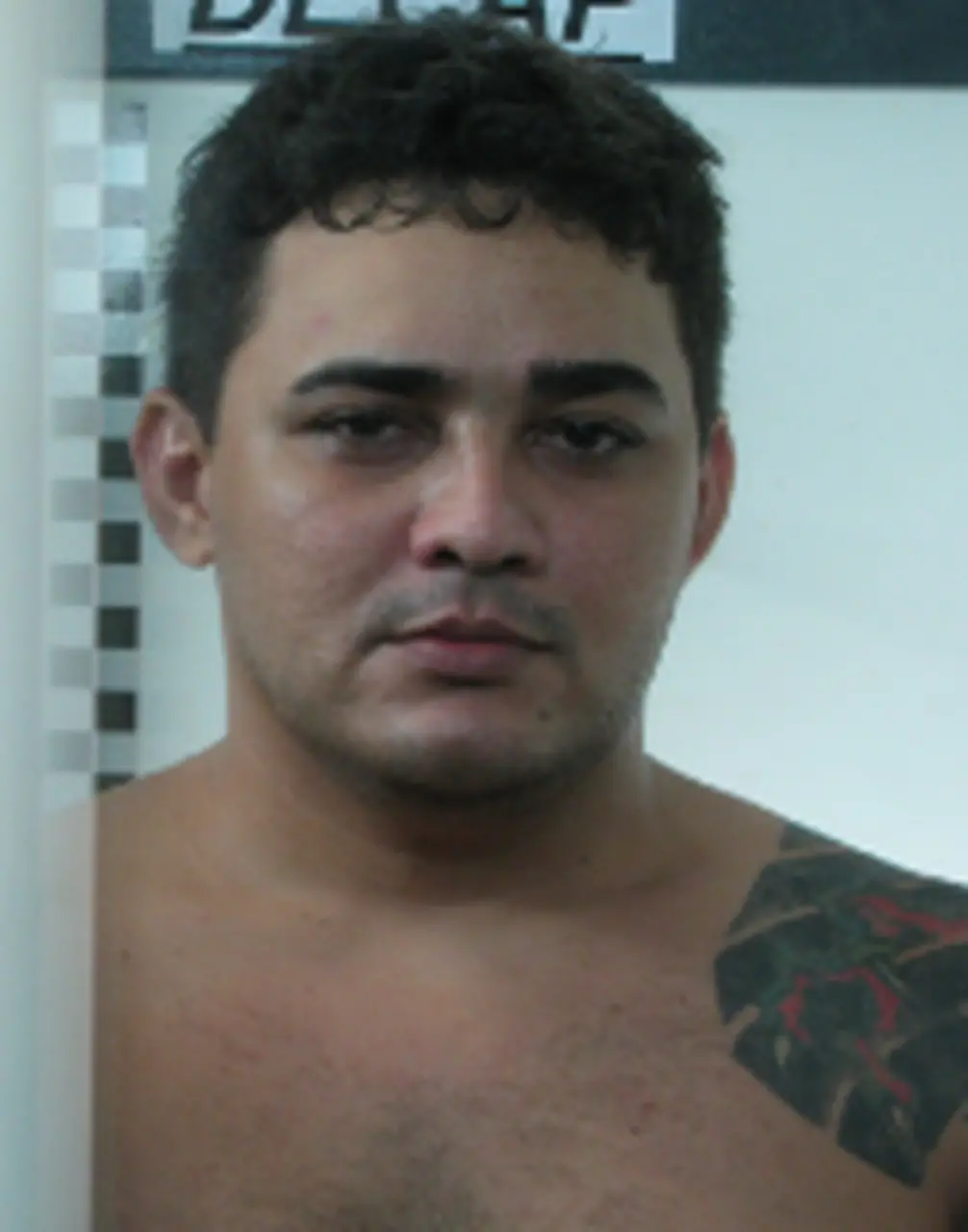 Imagem de um jovem rapaz com expressão neutra, usando camisa escura, com fundo branco e detalhes em cinza, em um ambiente interno bem iluminado.