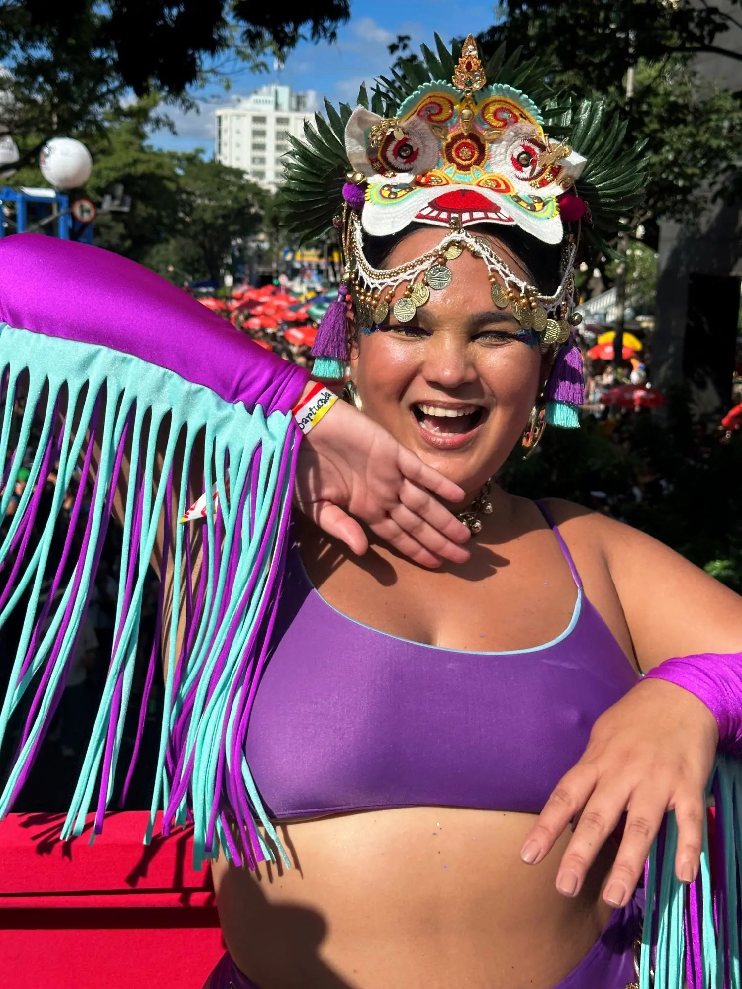 Mulher sorridente com cocar ornamentado e traje de carnaval roxo e turquesa, celebrando em um desfile ao ar livre.