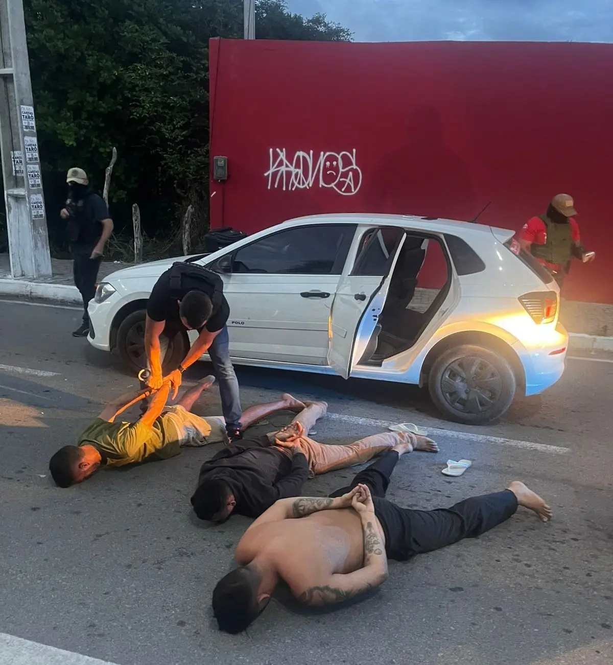 A imagem mostra três policiais, com coletes balísticos, realizando a prisão de três homens, que foram deitados no chão de uma via, após saírem de um carro branco.