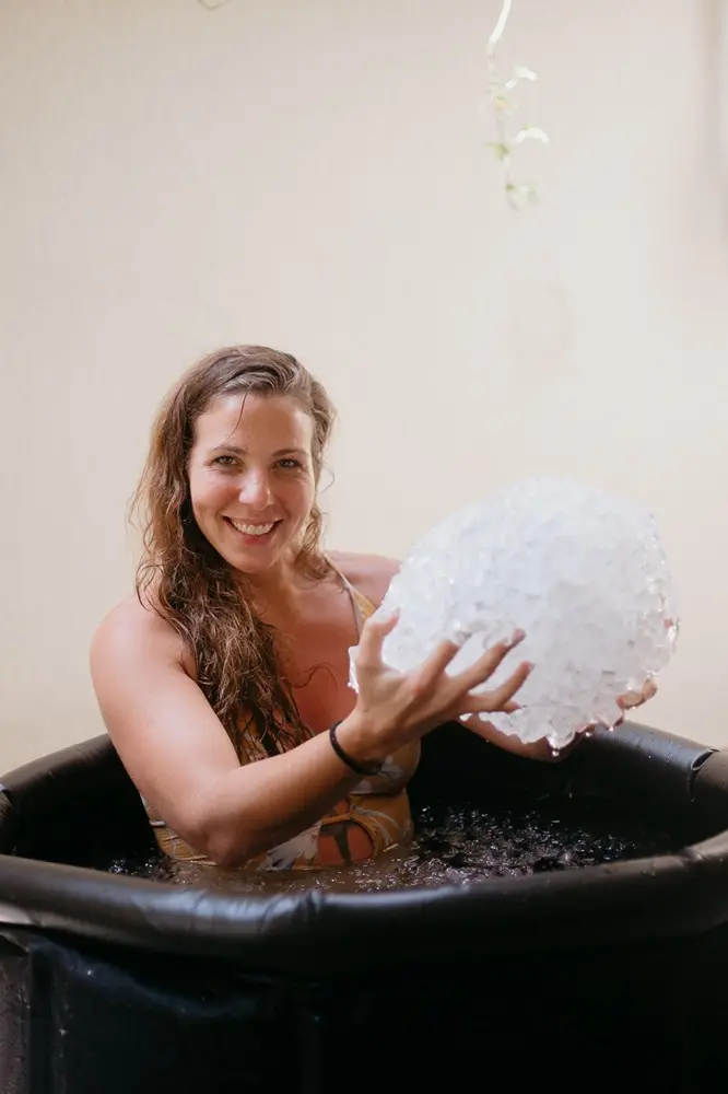 Na imagem, uma mulher sorridente com cabelos castanhos ondulados e úmidos está sentada dentro de uma banheira de imersão portátil preta. Ela segura com as duas mãos um grande bloco de gelo triturado à sua frente. Ela veste um traje de banho estampado em tons de amarelo e bege. O fundo é uma parede de tom neutro e claro, com uma pequena planta pendendo do topo.