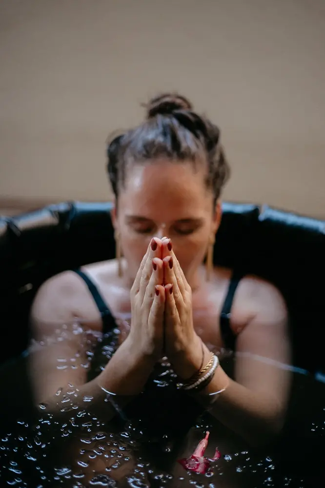 Na imagem, fotografia em close-up de uma mulher imersa em um banho de gelo, com foco nítido em suas mãos postas em posição de prece ou meditação à frente do rosto. Ela está com os olhos fechados, transmitindo uma expressão de concentração e serenidade. A água escura reflete gotas e pequenas bolhas na superfície, onde flutua uma única pétala de flor rosa. A mulher tem cabelos escuros presos em um coque, usa brincos dourados longos, pulseiras e esmalte vermelho escuro. O fundo está desfocado em tons de bege.