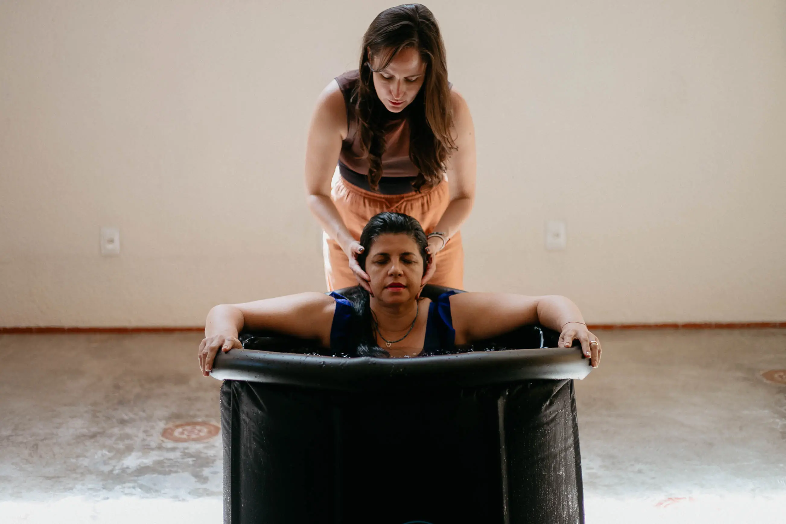 Na imagem, uma mulher de cabelos escuros está imersa até os ombros em uma banheira de gelo preta e cilíndrica. Ela está com os olhos fechados e expressão serena. Atrás dela, outra mulher, de pé, segura delicadamente as laterais do rosto da mulher imersa, oferecendo suporte. O ambiente tem paredes claras e chão de cimento, sugerindo um espaço de terapia ou bem-estar.