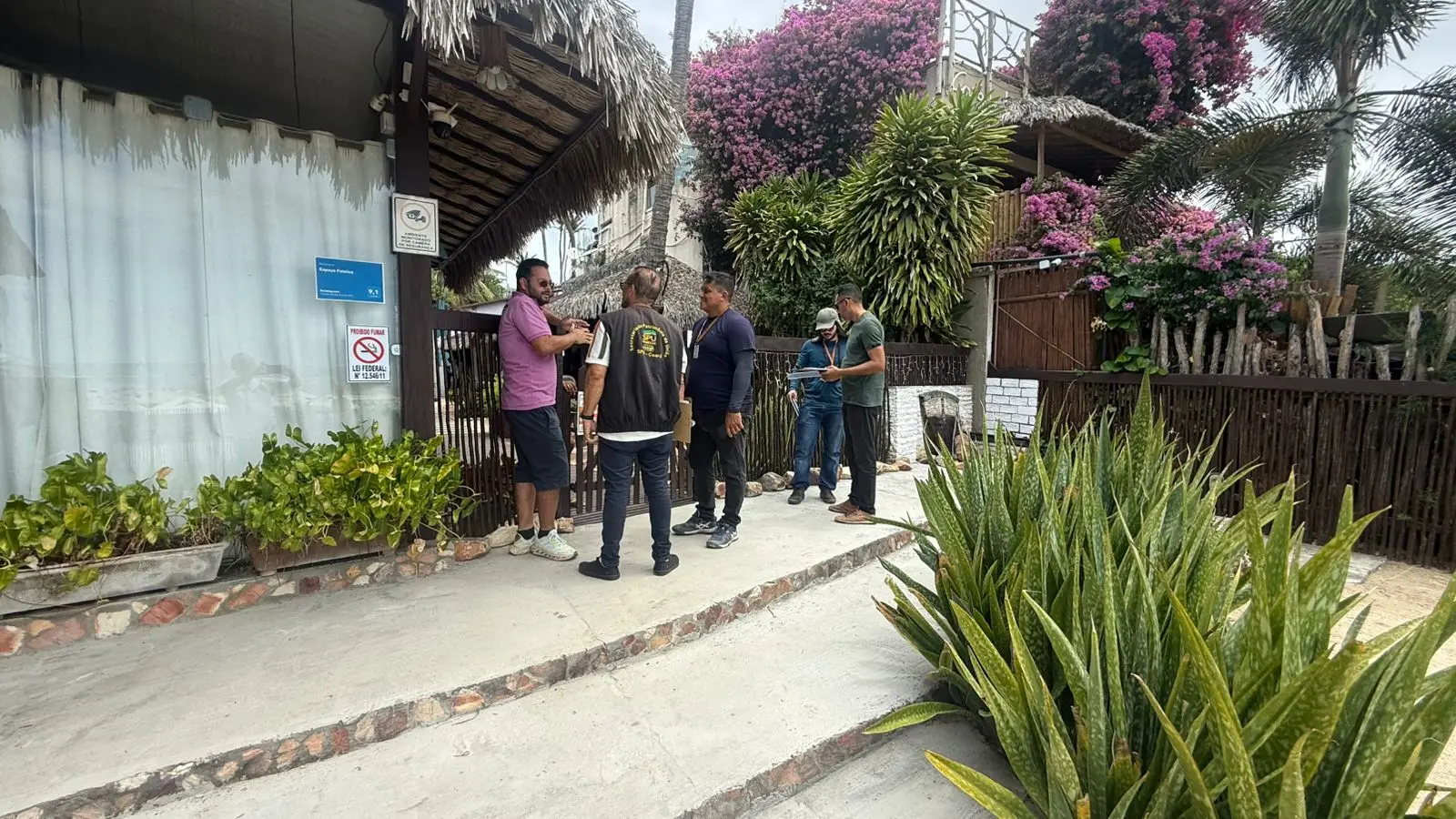 Um grupo de homens se reúne em frente a uma construção rústica com telhado de palha, cercada por plantas tropicais exuberantes e coloridas bougainvilleas roxas.
