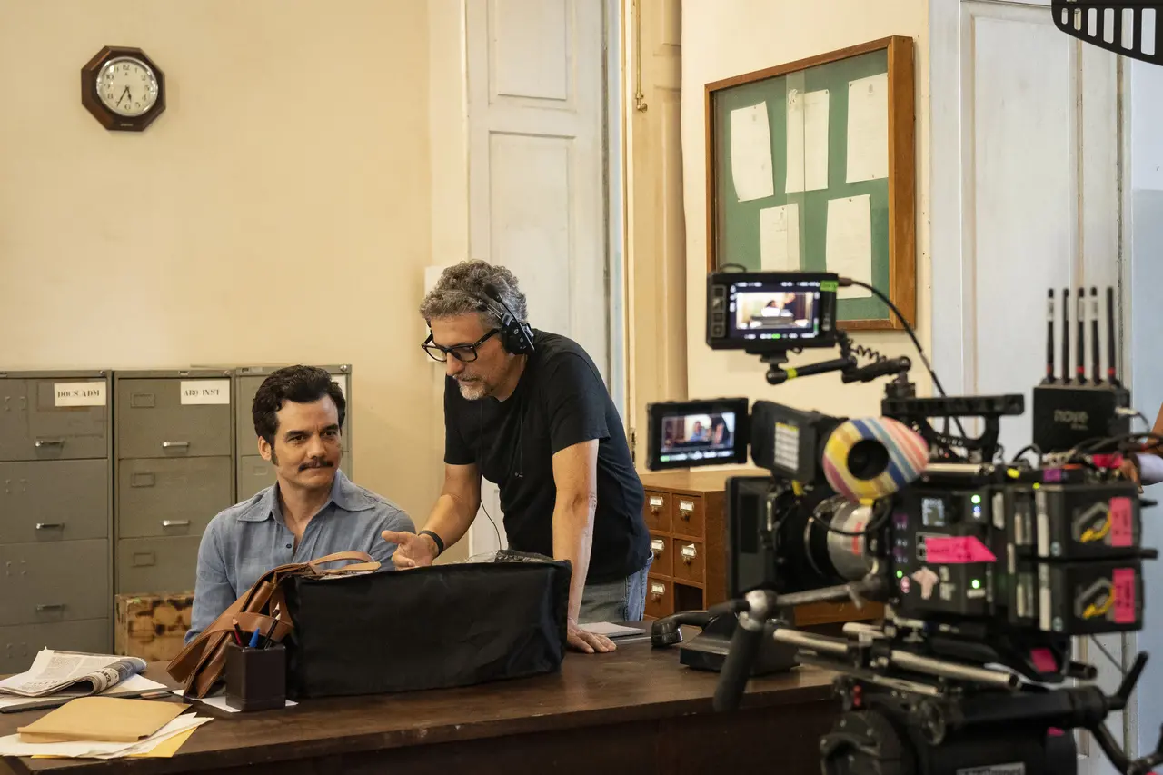 O ator Wagner Moura e o diretor Kleber Mendonça Filho conversam nos bastidores de um set de filmagem, sentados à frente de uma mesa de escritório antiga com arquivos de metal ao fundo. O enquadramento em plano médio destaca as câmeras de cinema em primeiro plano, capturando a colaboração entre os dois em um ambiente de estética vintage e profissional.