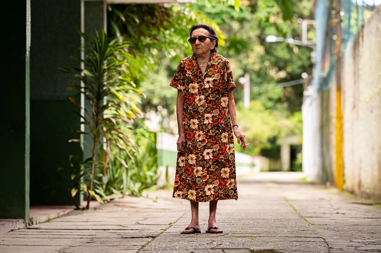 A atriz Tânia Maria posa em pé no centro de uma calçada arborizada, vestindo um vestido floral em tons de laranja e marrom enquanto segura um cigarro. A composição foca na personagem sob uma iluminação suave de dia, destacando sua expressão serena em meio ao cenário urbano com vegetação ao fundo.