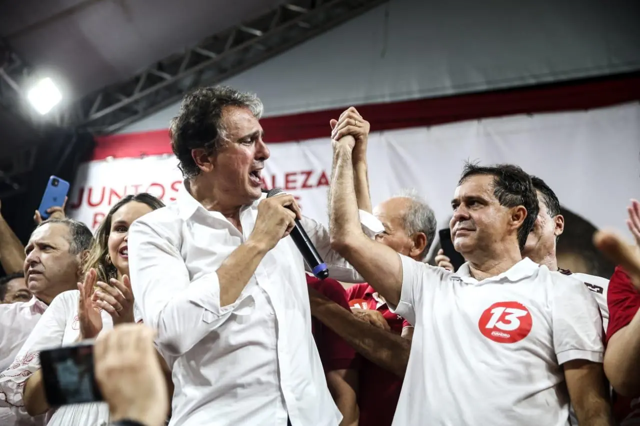 Camilo Santana fala ao microfone em um palco durante ato político, enquanto ergue a mão de Evandro Leitão  ao seu lado; ambos usam camisas claras, cercados por apoiadores, aplausos e celulares levantados, com banner ao fundo.