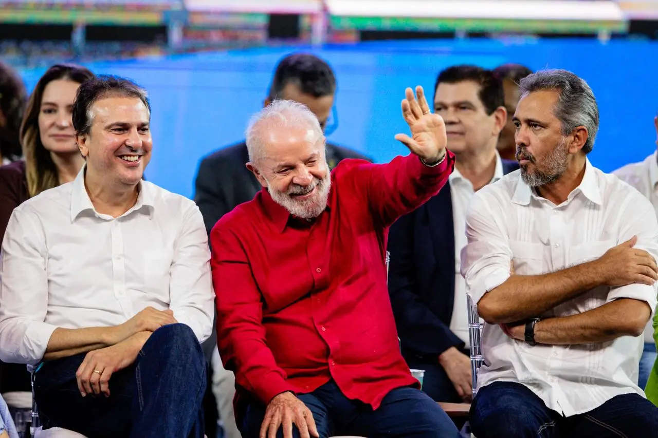 Camilo, Lula e Elmano sentados em um palco durante evento público; Lula ao centro, de camisa vermelha, sorri e acena para o público, enquanto os outros dois, de camisas claras, observam. Ao fundo, mais pessoas sentadas e um painel azul iluminado.