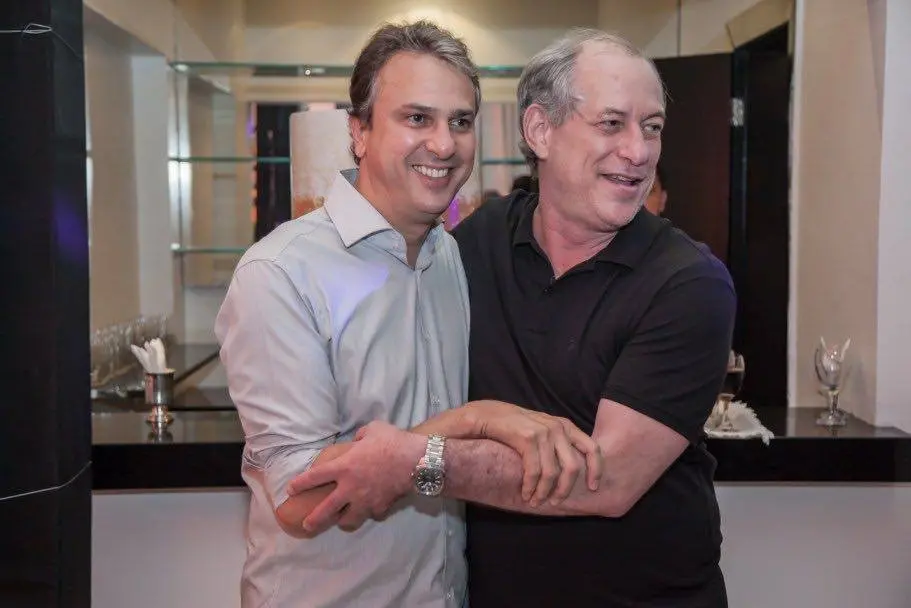 Camilo Santana e Ciro Gomes sorrindo em ambiente interno, de braços entrelaçados em gesto de confraternização. Camilo veste camisa clara de manga comprida e o Ciro usa camisa polo preta. Ao fundo, balcão com copos e prateleiras iluminadas.