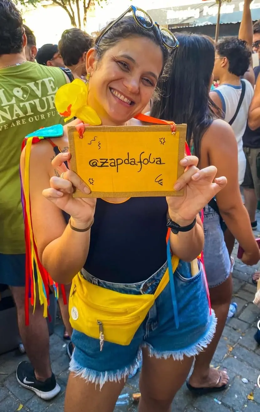 Na imagem, uma mulher jovem e sorridente posa em meio a uma multidão ao ar livre, possivelmente em um bloco de Carnaval. Ela tem pele clara, cabelos escuros presos e usa óculos de sol sobre a cabeça. Veste uma regata preta, shorts jeans de barra desfiada e uma pochete amarela vibrante na cintura. Em seus ombros, há adereços de fitas coloridas (arco-íris). Ela segura, com as duas mãos à frente do corpo, uma placa de papelão amarelo com o texto escrito à mão: 