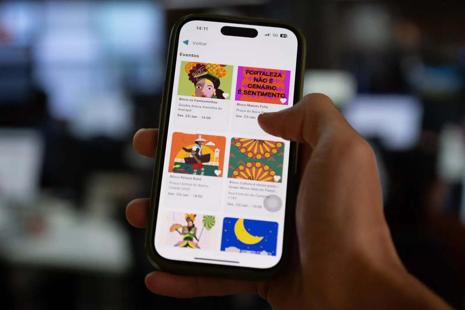 Na imagem, uma mão segura um smartphone (iPhone) que exibe a interface de um aplicativo de eventos culturais chamado 