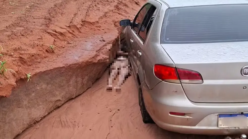 Carro prata parado em uma estrada de terra com um desnível profundo ao lado. Uma pessoa está caída no chão, parcialmente presa entre o carro e a parede de terra do barranco. O local aparenta ser uma área rural, com solo avermelhado e sinais de erosão.