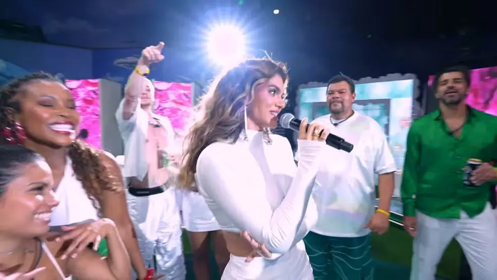 Na imagem, uma fotografia colorida e vibrante capturada em um ângulo levemente baixo, mostrando a cantora Kelly Key se apresentando em um palco ou cenário de festa. Ela está de perfil para a direita, segurando um microfone próximo à boca, com um leve sorriso. Ela tem cabelos longos, ondulados e loiros, e veste um conjunto branco de manga comprida e gola alta. Ao redor dela, várias pessoas celebram com entusiasmo.