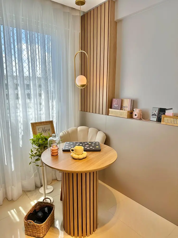 Na imagem, um canto de escritório decorado em tons pastéis e madeira clara. No centro, uma mesa redonda ripada com um notebook adesivado, a xícara amarela e a vela acesa. Atrás da mesa, uma cadeira estofada em formato de concha. O cenário inclui uma cortina branca translúcida, uma planta em um pedestal e uma prateleira lateral com itens decorativos como cadernos, uma câmera instantânea rosa e pequenos quadros. Uma luminária pendente dourada com globo branco ilumina o espaço.