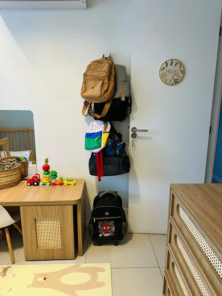 Na imagem, vista de um quarto infantil organizado. Penduradas em ganchos na parede branca, estão várias mochilas de diferentes tamanhos e cores. No chão, uma mochila de rodinhas do Homem-Aranha. À esquerda, uma bancada de madeira com brinquedos de montar coloridos e um cesto de palha. À direita, uma cômoda de madeira com gavetas de palha trançada. Na porta branca ao centro, há um enfeite circular personalizado com o nome 