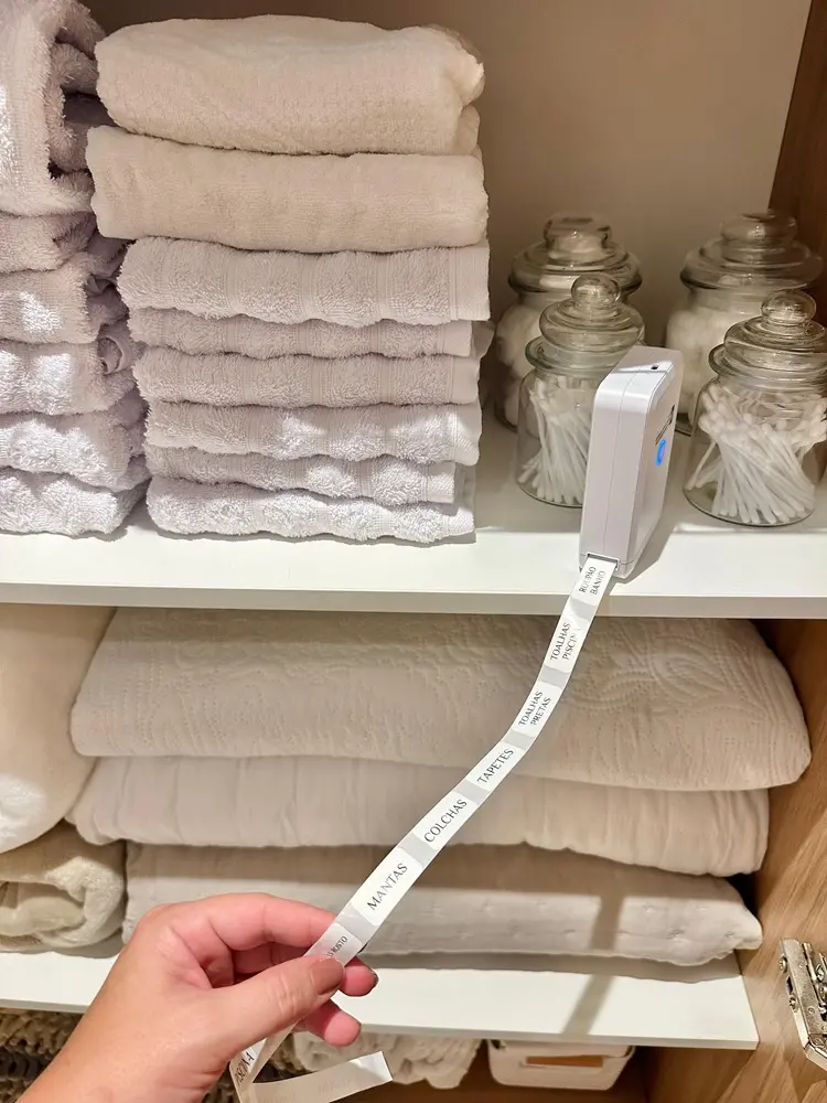 Na imagem, uma visão em close do interior de um armário de roupas de banho branco, organizado de forma impecável. No lado esquerdo, há pilhas de toalhas brancas e lilás claro dobradas. À direita, três potes de vidro redondos contêm itens de higiene como algodão e hastes flexíveis. No centro da prateleira, um rotulador eletrônico branco e compacto está imprimindo uma fita de etiquetas adesivas. Uma mão segura a extremidade da fita, que mostra palavras impressas em português como 