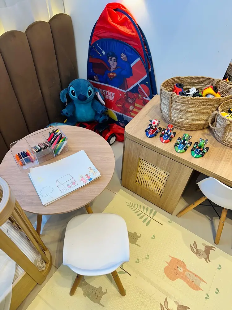Na imagem, uma área de lazer infantil organizada e iluminada. No centro esquerdo, há uma pequena mesa redonda de madeira com um caderno aberto exibindo um desenho infantil de uma casa e pessoas, acompanhado de um organizador de acrílico com giz de cera e lápis coloridos. À frente da mesa, uma pequena cadeira branca. Ao lado, um móvel de madeira clara serve de bancada para quatro carrinhos de brinquedo do Mario Kart alinhados e um cesto de palha com outros brinquedos. Ao fundo, vê-se um boneco de pelúcia do Stitch sentado próximo a uma barraca temática da Liga da Justiça (Superman e Flash). O chão é coberto por um tapete emborrachado bege com ilustrações suaves de animais e folhas.
