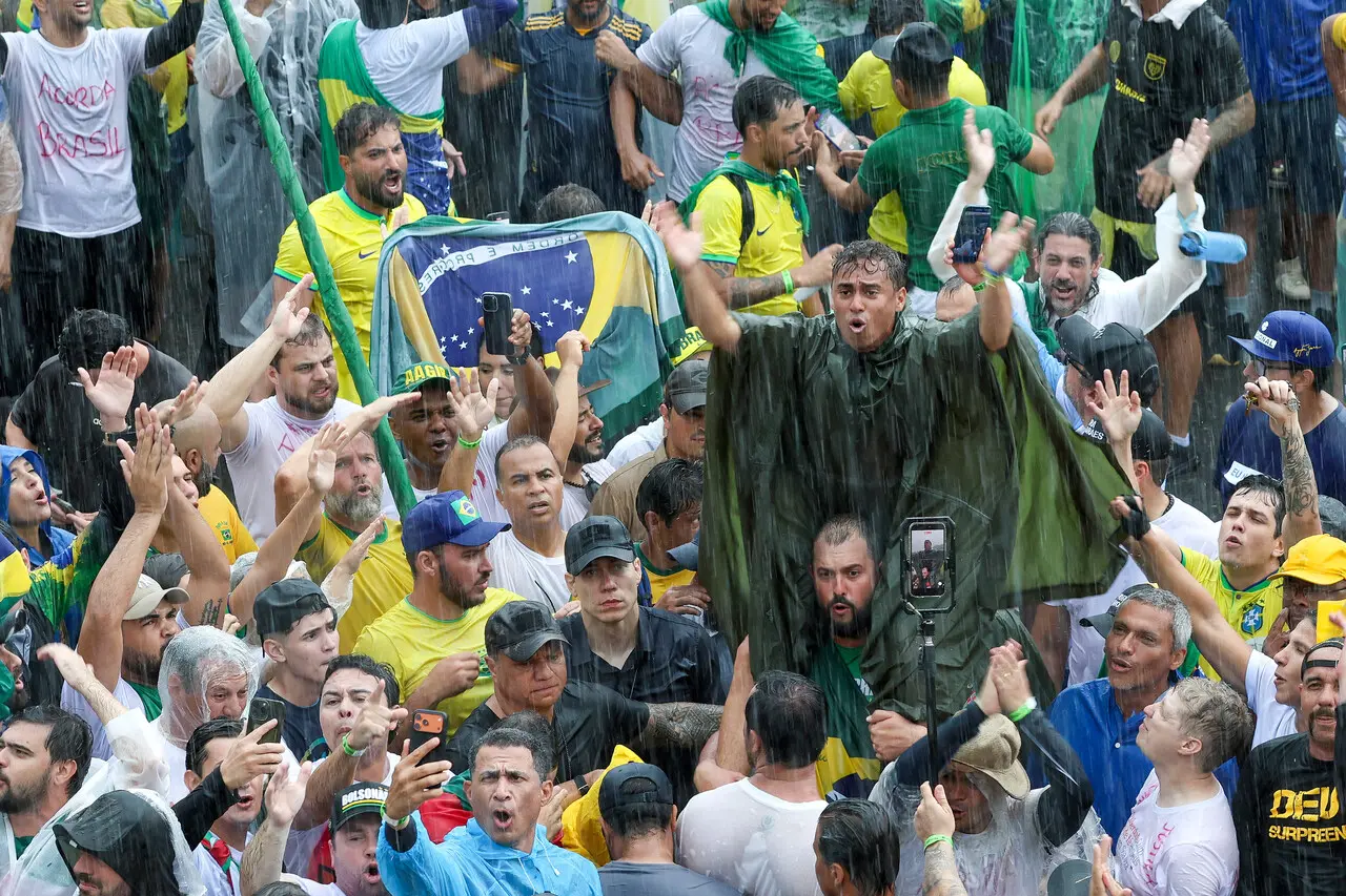 Multidão reunida sob forte chuva em manifestação com bandeiras do Brasil, roupas verde e amarelas, camisetas com frases políticas e pessoas erguendo os braços enquanto registram o momento com celulares.