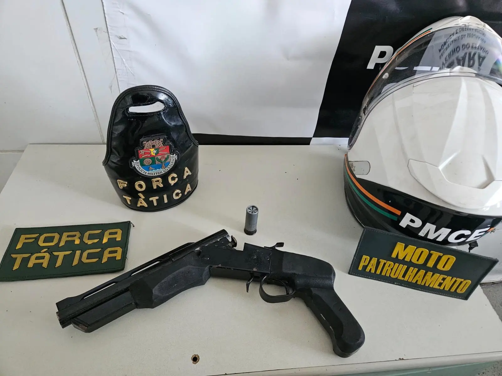 Imagem mostra uma espingarda caseira apreendida pela polícia, ao lado de capacete e distintivo da polícia.