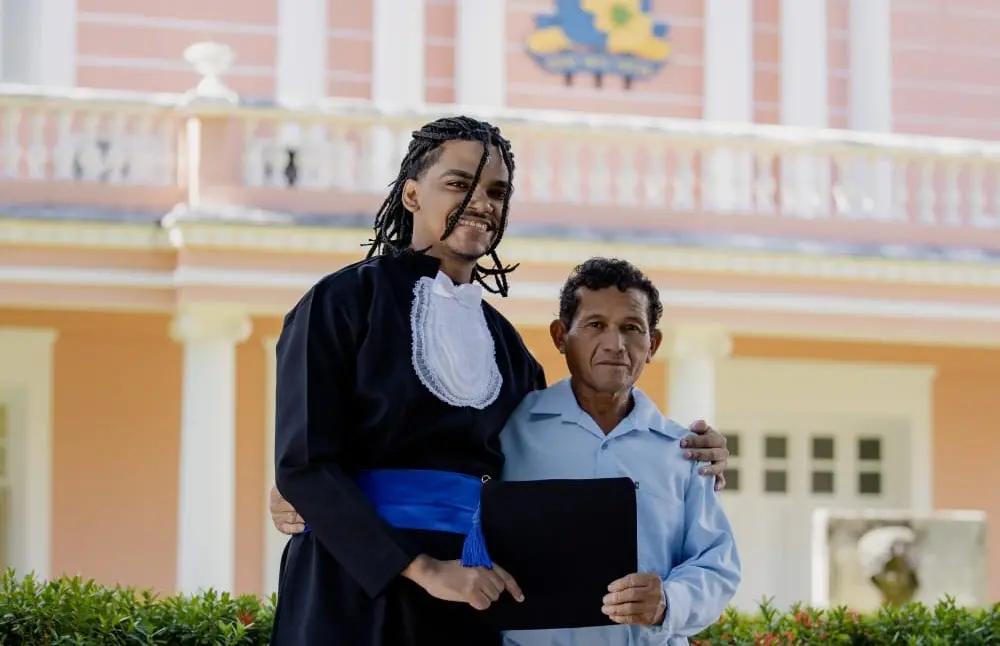 Emanuel Ferreira sorridente com cabelo dread, vestindo toga de formatura, ao lado de um senhor com camisa azul, em frente a uma residência colorida durante uma cerimônia de formatura na Universidade Federal do Ceará (UFC).