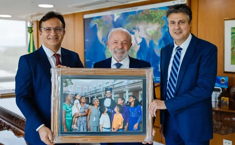 Lula, Custódio e Camilo apresentando uma foto de Emanuel Ferreira e família na sala de reuniões, com um mapa-múndi ao fundo. Evento relacionado à política ou educação.