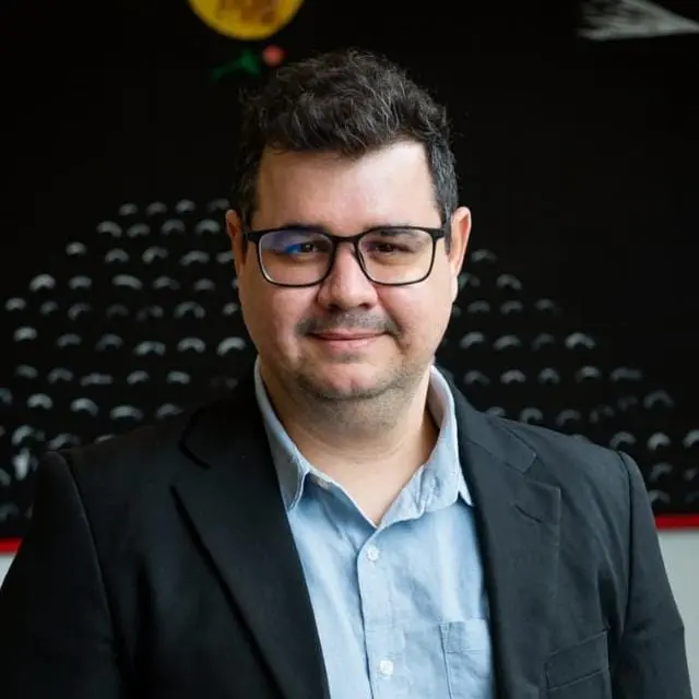 Homem de óculos e cabelo curto escuro, vestindo camisa azul clara e blazer preto, posa para retrato em ambiente interno com fundo escuro e elementos gráficos desfocados.