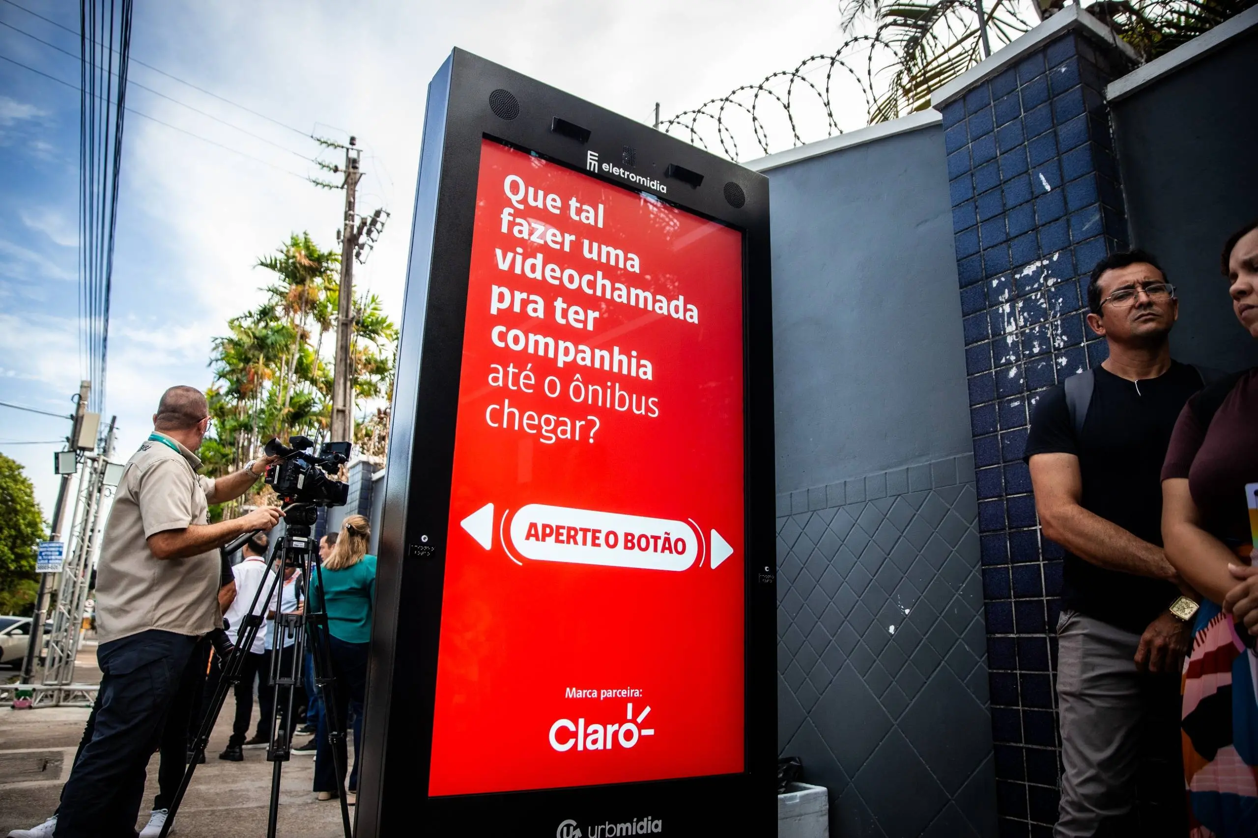 Painel digital de ponto de ônibus exibindo anúncio da Claro com mensagem sobre fazer videochamada enquanto espera o transporte, instalado em área urbana com jornalistas e câmeras ao fundo.