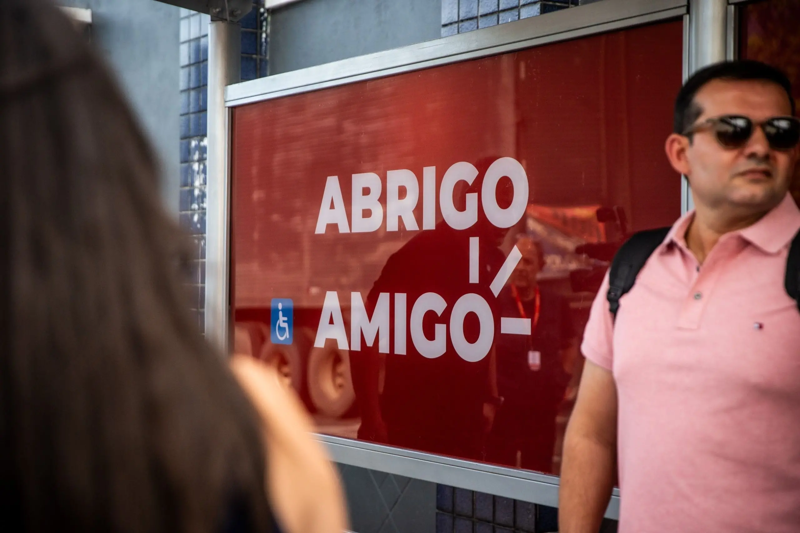 Abrigo de ponto de ônibus com painel vermelho exibindo a frase 'Abrigo Amigo', com pessoas próximas ao local e ambientação urbana ao redor.