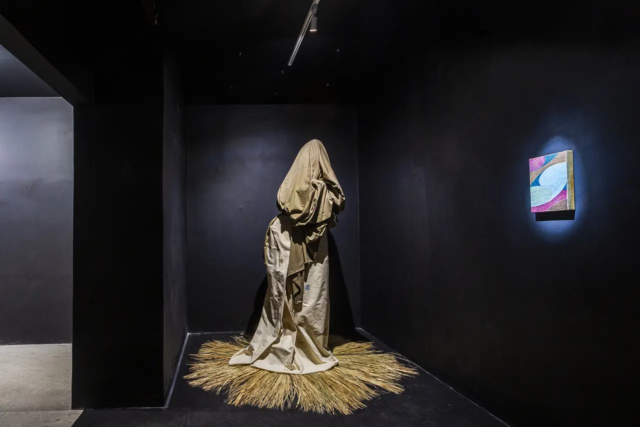 Uma obra de arte envolta em tecidos beges e tons terrosos ocupa o centro de uma sala escura na exposição 