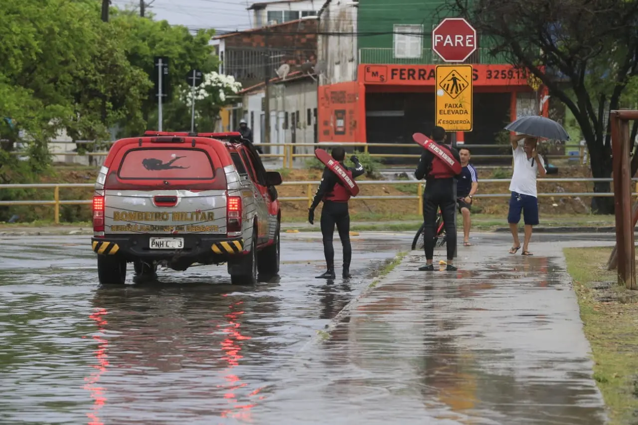 Cena de alagamento em via urbana. Um veículo do Corpo de Bombeiros está parado sobre a rua inundada, enquanto dois bombeiros caminham ao lado com flutuadores vermelhos. Ao fundo, duas pessoas seguram um guarda‑chuva na calçada molhada.
