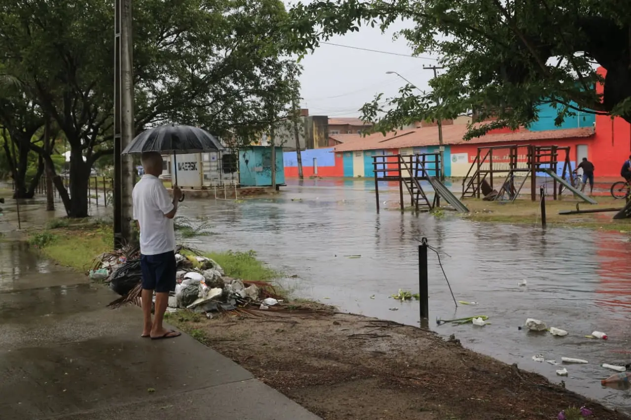 Homem com guarda-chuva observa canal em bairro residencial, com playground submerso e casas coloridas ao fundo.
