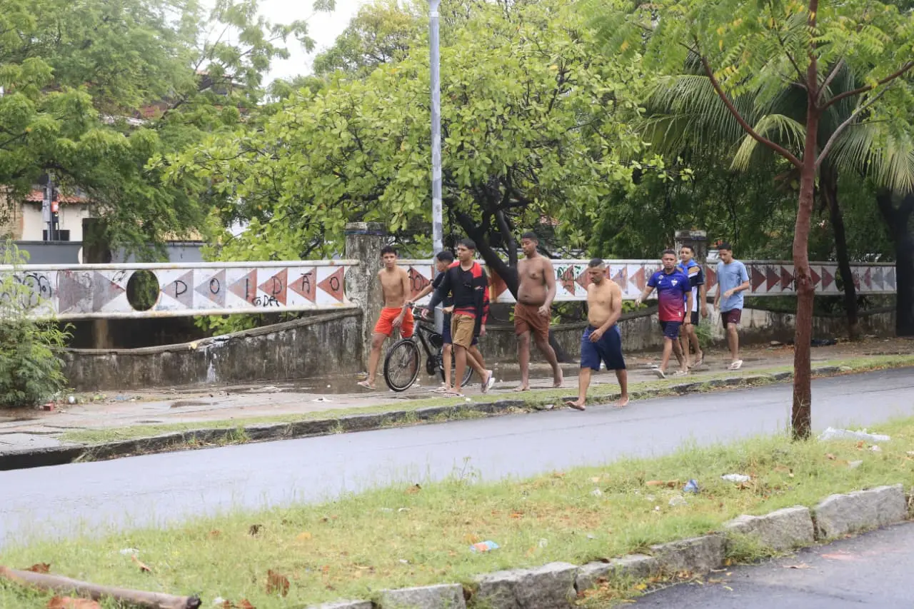 Grupo de jovens, alguns sem camisa e descalços, caminhando na beira de uma estrada molhada com uma bicicleta, tendo uma ponte e árvores ao fundo.