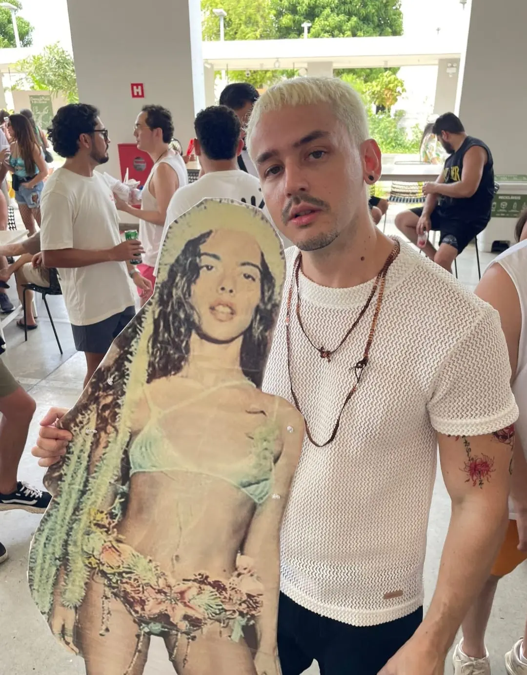 Homem que se fantasiou de Juliano Floss, com cabelo loiro platinado e camiseta branca texturizada, encara a câmera enquanto segura um totem de papelão da cantora Marina Sena. Ele está em um local aberto com outras pessoas ao fundo e usa colares de contas. O totem mostra a artista em um traje de banho verde claro, com uma planta de cacto ao lado.