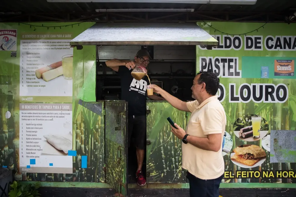 Na imagem, fotografia colorida em plano médio mostra a fachada de um quiosque de rua verde vibrante, especializado em caldo de cana e lanches. No centro, um atendente homem, usando boné, óculos e camiseta preta, está inclinado para fora de uma pequena janela, servindo caldo de cana de uma jarra em um copo plástico. Um cliente, um homem de pele clara vestindo uma camisa polo bege, está de pé à direita, estendendo a mão para receber a bebida enquanto segura um celular na outra mão. O quiosque é decorado com grandes adesivos informativos. À esquerda, há uma lista intitulada 