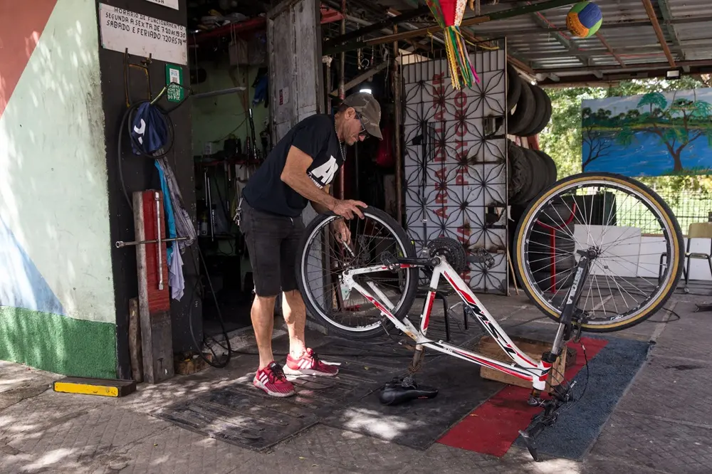Na imagem, um homem de boné, camiseta preta e bermuda jeans trabalha no conserto de uma bicicleta branca e vermelha, que está posicionada de cabeça para baixo sobre um tapete de borracha. Ele está inclinado, manuseando a roda traseira em uma oficina rústica e aberta (borracharia). Ao fundo, veem-se pneus empilhados, ferramentas e uma parede com uma pintura de paisagem. No canto superior esquerdo, há uma placa com o horário de funcionamento escrito à mão. A iluminação é natural, sugerindo um ambiente externo coberto.