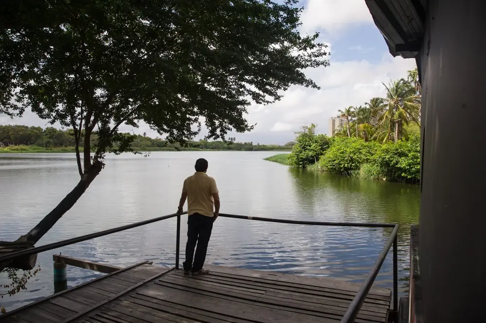 Na imagem, uma fotografia em plano médio mostra um homem de pele morena, vestindo uma camisa polo amarela clara e calças escuras, posicionado de costas no centro de um deck de madeira rústica. Ele está apoiado em um corrimão de metal preto, olhando para uma vasta extensão de água (uma lagoa ou rio largo). À esquerda, uma árvore de copa densa projeta sombra sobre o deck. À direita, a margem é composta por vegetação verde exuberante e vários coqueiros. Ao fundo, sob um céu com nuvens brancas e cinzas, nota-se a silhueta de um edifício distante. A cena transmite uma sensação de tranquilidade e contemplação.