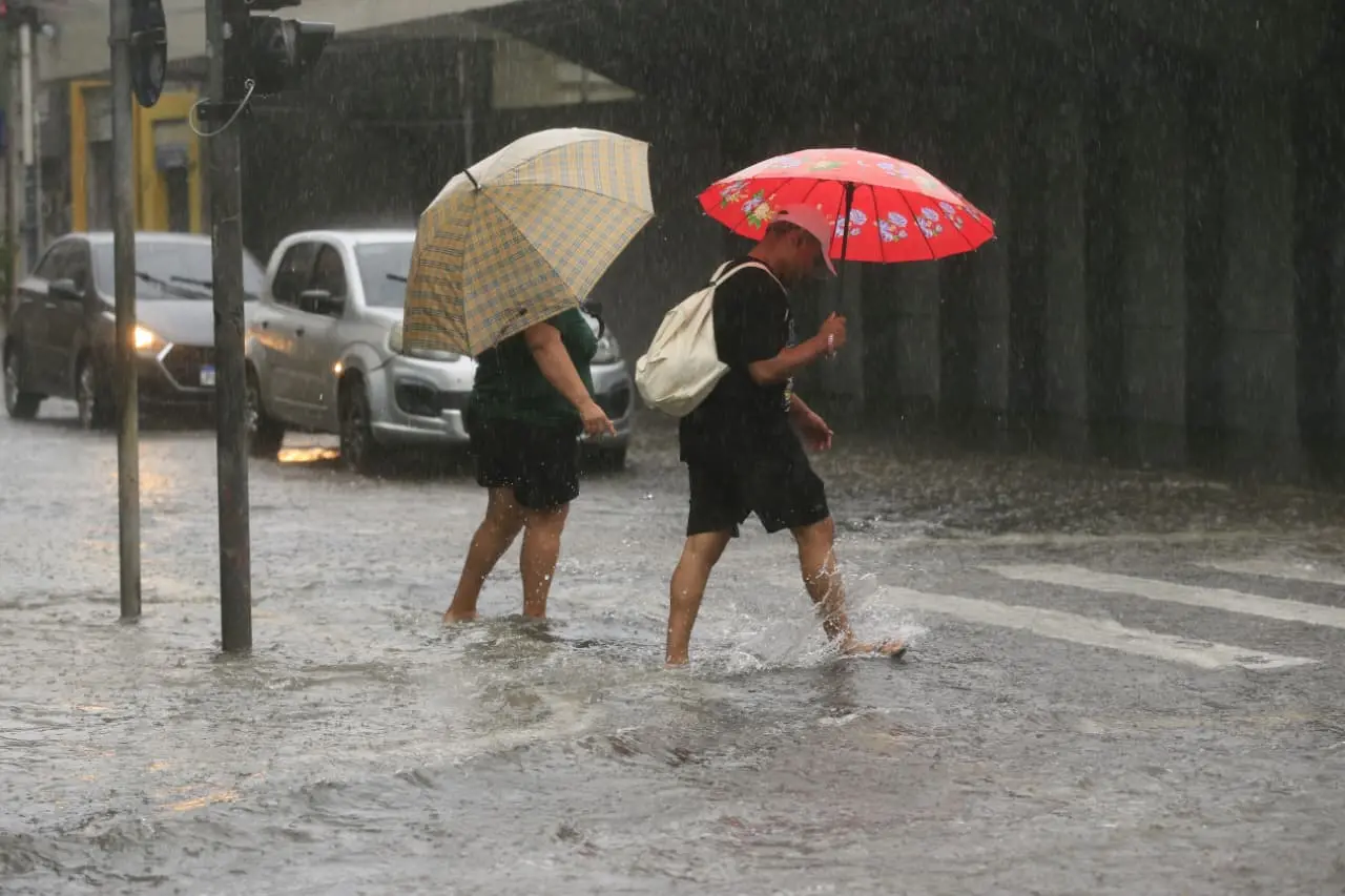 Imagem mostra duas pessoas atravessando uma rua alagada.