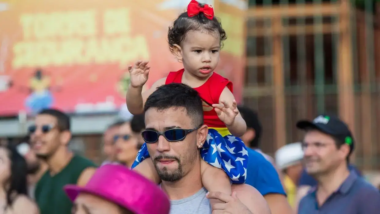 Um homem com óculos escuros carrega uma criança pequena vestindo uma fantasia de Mulher Maravilha nos ombros.