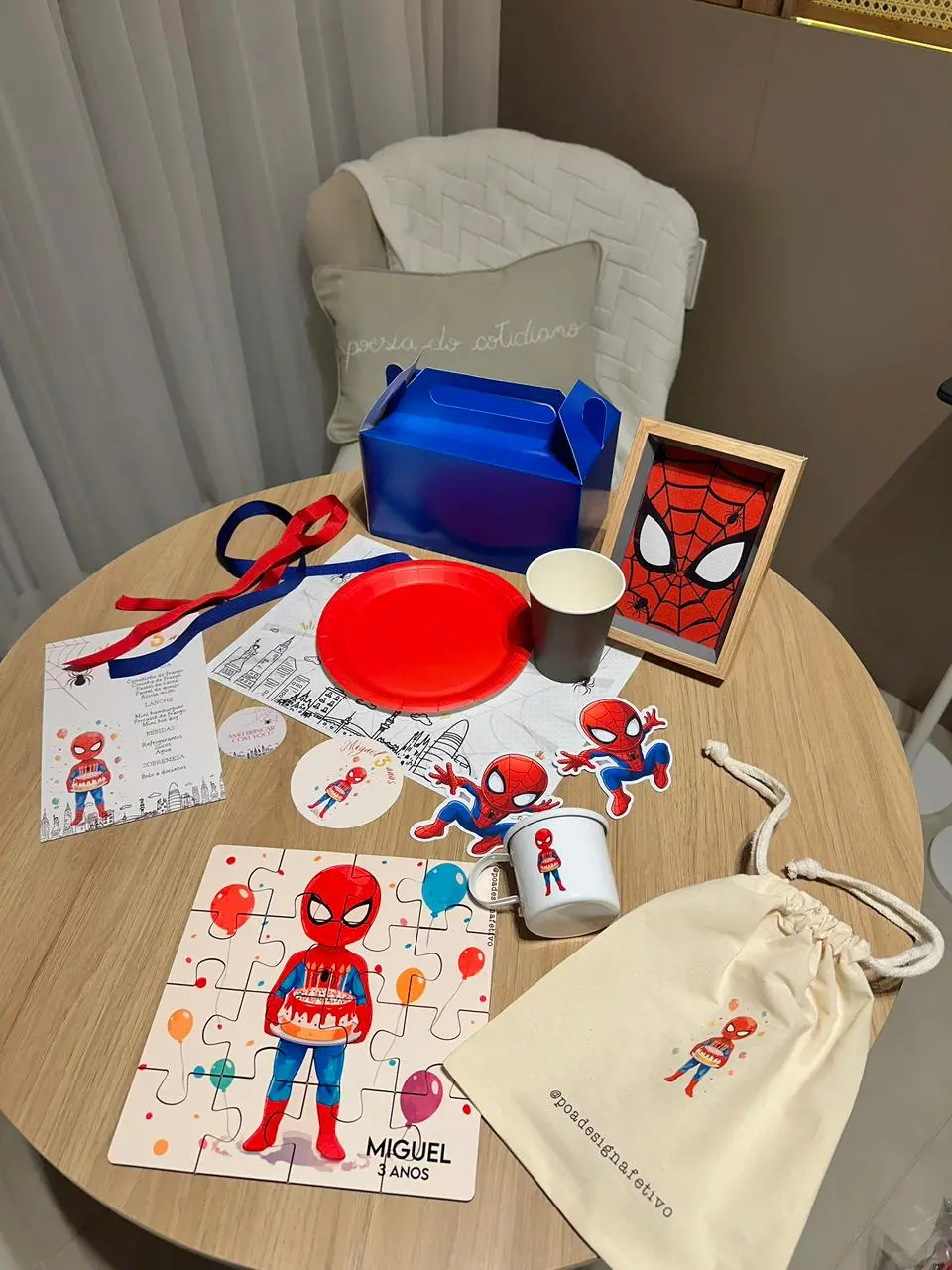 Na imagem, vista superior de uma mesa redonda de madeira clara organizada com itens de uma festa infantil com o tema Homem-Aranha. No centro da mesa, há um prato de papel vermelho, um copo preto e uma caixa de lanche azul metálica. À esquerda, um menu personalizado com o nome 