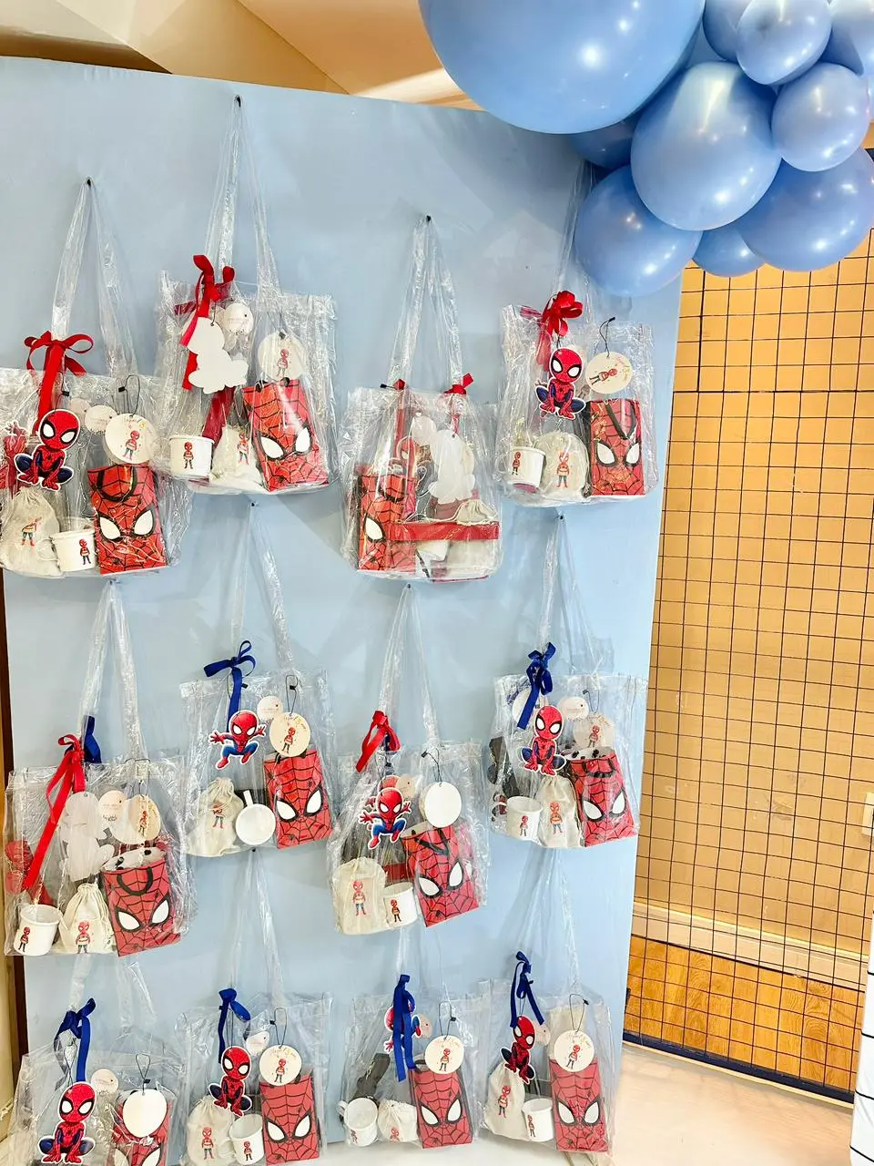Na imagem, várias sacolas de lembrancinhas transparentes com o tema do Homem-Aranha estão penduradas em um painel azul claro em uma festa infantil. Aproximadamente doze sacolas de plástico transparente estão dispostas em fileiras, penduradas por alças finas em um painel vertical azul claro.