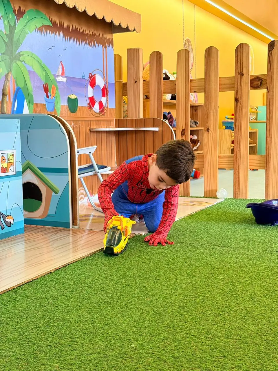 Na imagem, um menino vestido de Homem-Aranha brinca com um carrinho amarelo sobre um gramado sintético em uma área de recreação. A criança está ajoelhada no centro da imagem, vestindo uma fantasia completa do Homem-Aranha. Ela está concentrada, empurrando um brinquedo amarelo que parece um veículo ou helicóptero.