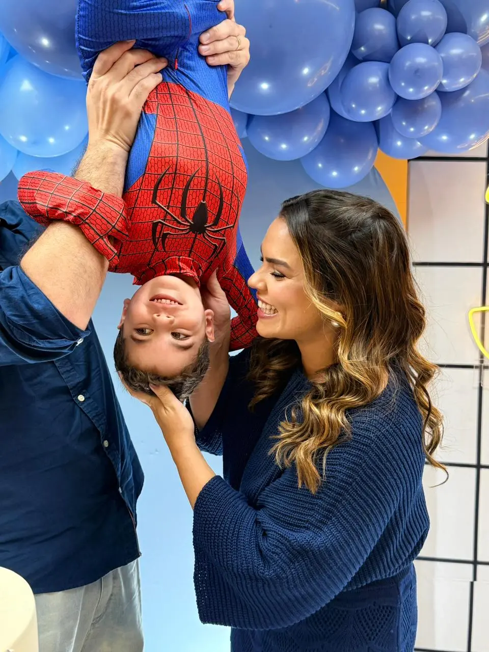 Na imagem, uma fotografia vertical em um ambiente de festa. Um homem (visto parcialmente de lado) segura uma criança de ponta-cabeça, imitando a clássica pose do Homem-Aranha. A criança, um menino, veste uma fantasia do herói e sorri para a câmera. À direita, uma mulher com cabelos castanhos ondulados e um suéter azul marinho segura delicadamente a cabeça do menino enquanto sorri com ternura para ele. O fundo é decorado com balões azuis e uma parede quadriculada branca.