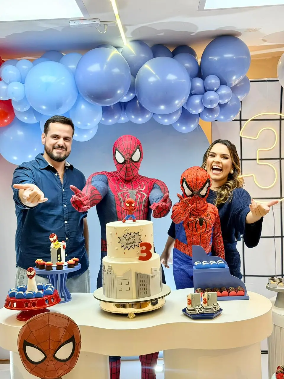Na imagem, uma fotografia colorida de uma festa de aniversário de 3 anos com o tema Homem-Aranha. No centro, atrás de uma mesa branca ondulada, estão um adulto fantasiado de Homem-Aranha e uma criança pequena, também com a fantasia completa e máscara, ambos fazendo o gesto de 