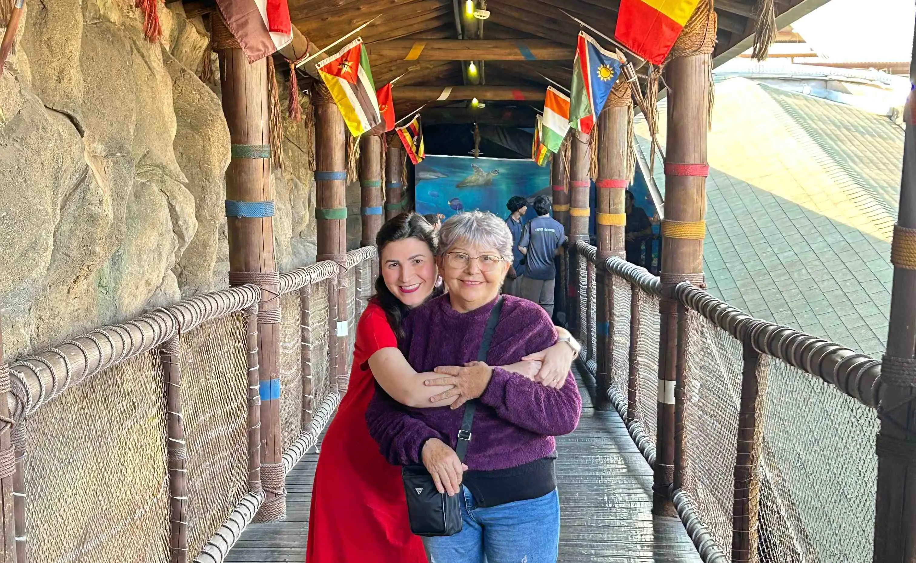 Franciele e Liduína se abraçam e sorriem em uma passarela rústica decorada com bandeiras de vários países. Franciele veste vermelho e Liduína usa blusa roxa, óculos e uma bolsa transversal.
